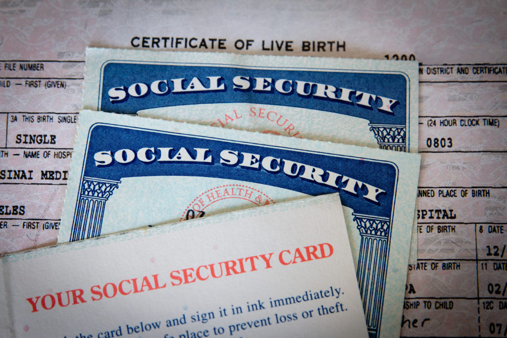 wo USA Social Security cards with number obscured surrounded by US dollars or American dollars. The SSN has become a de facto national identification number for taxation and other purposes. United States Social Security cards with birth certificate background. Concept for life, retirement or death or Government documents and identification.
